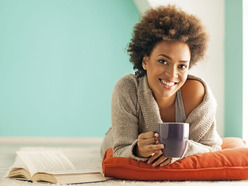 smiling woman lying on cushion on the floor and holding a cup of tea, with a book open beside her
