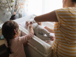 Little girl watching her mum change her baby sibling on a changing table in a bedroom
