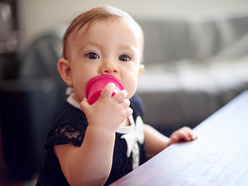 a baby with a toy in her mouth standing up and holding the edge of a coffee table 