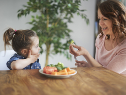 mum showing toddler daughter a broccoli floret from a plate of vegetables